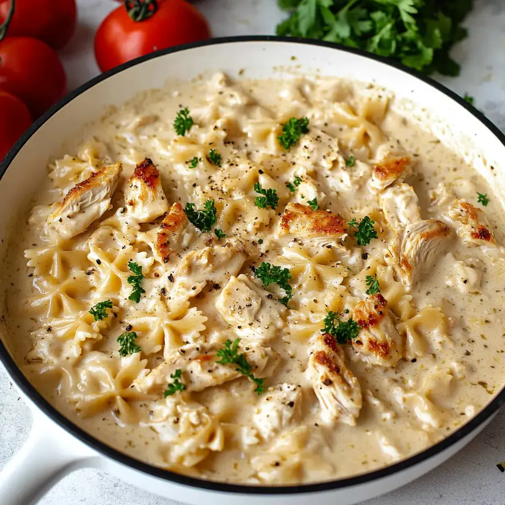 A creamy pasta dish with chunks of grilled chicken, garnished with parsley, served in a white skillet alongside fresh tomatoes and cilantro.