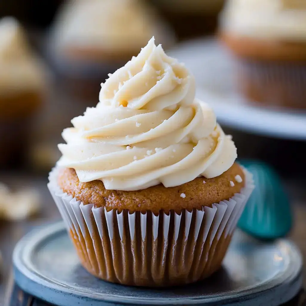 A close-up of a frosted cupcake with a swirl of creamy vanilla icing on top, placed on a metallic plate.