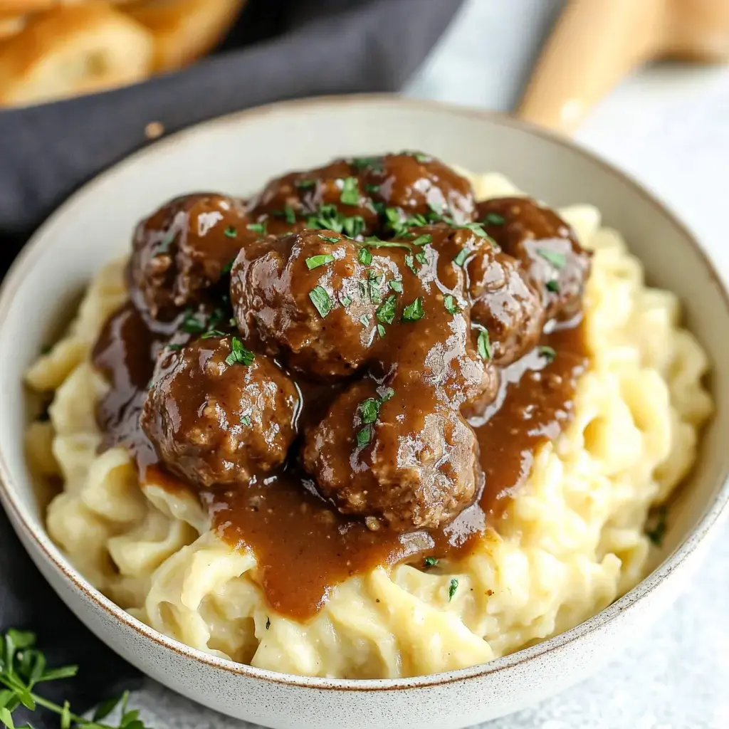 A bowl of creamy pasta topped with meatballs and brown gravy, garnished with chopped parsley.