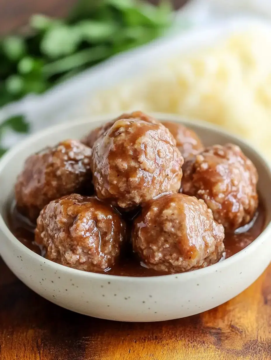 A bowl of meatballs in a savory brown sauce, garnished with fresh herbs in the background.