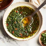 A bowl of vibrant green herb sauce with a spoon, surrounded by a small dish of fresh chopped herbs.