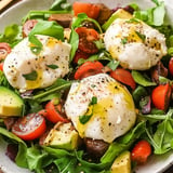 A vibrant salad featuring fresh greens, cherry tomatoes, avocado, olives, and three poached eggs drizzled with olive oil and sprinkled with black pepper and herbs.