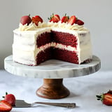 A red velvet cake with cream cheese frosting, topped with fresh strawberries, sits on a marble cake stand with a slice removed.