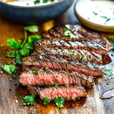 Sliced grilled steak garnished with fresh parsley, resting on a wooden cutting board, with a bowl of sauce in the background.