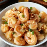 A close-up of a white bowl filled with shrimp topped with chopped green onions and nuts in a savory sauce.