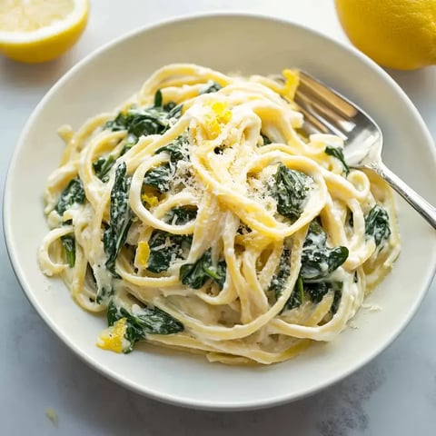 A serving of creamy fettuccine pasta with spinach and lemon, garnished with grated cheese, in a white bowl next to a lemon half.