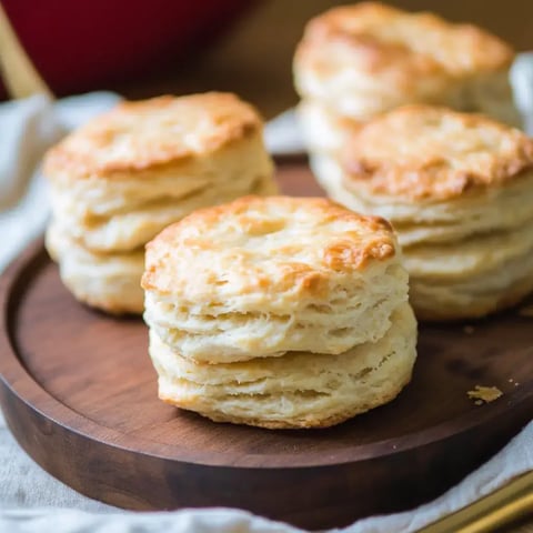 A wooden platter holds several golden-brown biscuits, showcasing their flaky layers.